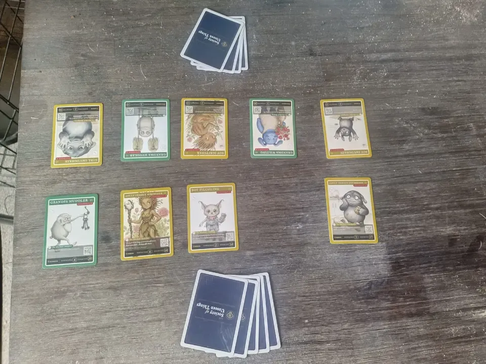 Nine printed catalogue cards of household creatures laid out on a table, with two stacks of the navy card backs.