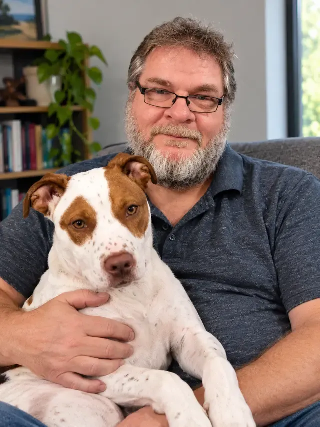 Dirk Wessels, bearded and in glasses, sitting on a couch at home with a brown-and-white dog in his arms.