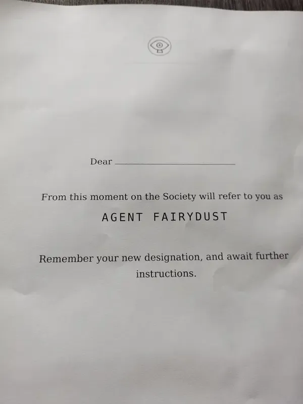 The first note from the Society, printed on A4: a small eye crest, 'Dear' and a blank line, then 'From this moment on the Society will refer to you as Agent FairyDust. Remember your new designation, and await further instructions.'
